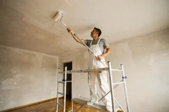 Peintre professionnel appliquant de la peinture blanche sur un plafond de 50m2 avec un rouleau à long manche depuis un échafaudage en aluminium